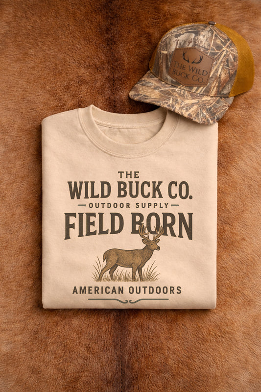 Beige t-shirt with 'The Wild Buck Co.' branding on a brown leather surface with a camouflage cap.